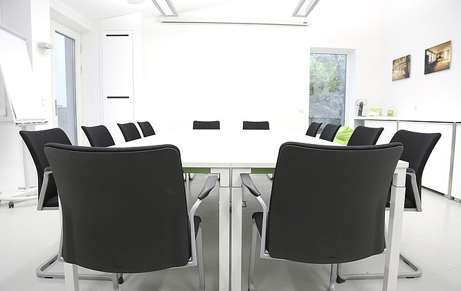 Unser Lounge - für Termine, Events und Mittagessen Heller, moderner Besprechungsraum mit großem weißen Konferenztisch, umgeben von schwarzen Stühlen, Flipchart, Fenster mit Tageslicht und minimalistischer Einrichtung.