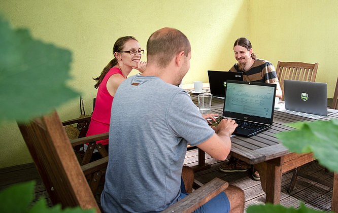 Drei Kolleginnen und Kollegen arbeiten gemeinsam mit Laptops an einem Holztisch im schattigen Gartenbüro; sie lachen, trinken Kaffee und analysieren Daten in entspannter Outdoor-Atmosphäre.
