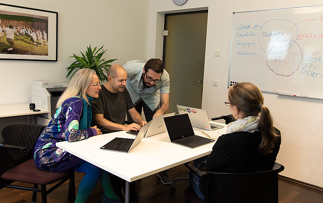Fünf Personen sitzen gemeinsam in einem modernen Besprechungsraum mit Laptops an einem Tisch und diskutieren konzentriert vor einem Whiteboard mit Marketing-Diagramm; an der Wand hängen ein Teamfoto und eine große Zimmerpflanze steht im Hintergrund.