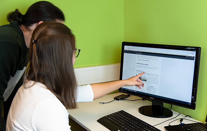 Zwei Mitarbeitende analysieren gemeinsam Inhalte auf einem großen Monitor in einem modernen Büro mit grüner Wand; eine Frau zeigt auf eine Website mit dem Titel „Google My Business – Die Grundlagen“.