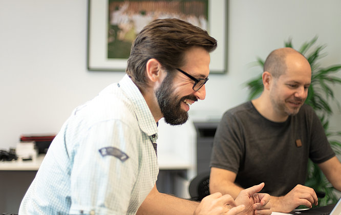 Zwei Männer lachen gemeinsam während eines Meetings in einem modernen Büro; einer arbeitet am Laptop, im Hintergrund sind eine große Zimmerpflanze und ein gerahmtes Foto an der Wand zu sehen.