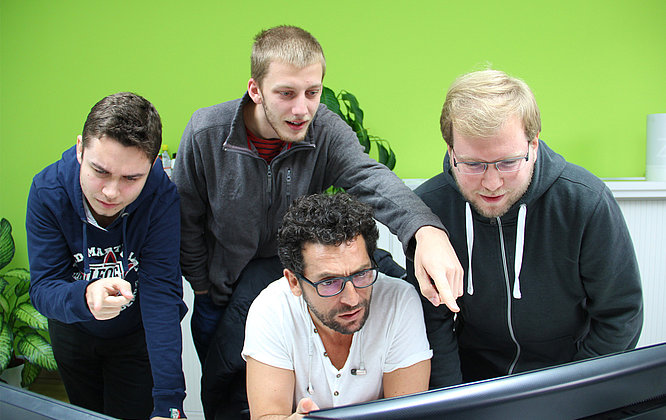 Ein TYPO3-Team Vier männliche Entwickler stehen und sitzen vor einem Computerbildschirm in einem modernen Büro mit grüner Wand und analysieren gemeinsam intensiv den Bildschirminhalt.