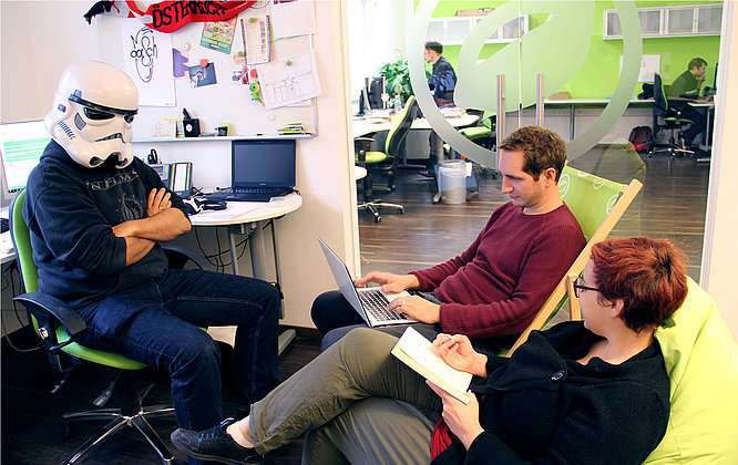 Erster Stock in Wien. Szene nicht ganz ernst zu nehmen :-) Drei Mitarbeitende halten ein informelles Meeting im Büro ab – eine Person trägt humorvoll einen Stormtrooper-Helm, während die anderen auf einem Sitzsack und mit Laptop diskutieren; im Hintergrund ist ein modernes, grünes Großraumbüro mit weiteren Arbeitsplätzen zu sehen.